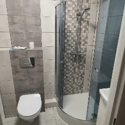 Nowoczesna łazienka z kabiną prysznicową narożną, toaletą wiszącą i szaro-białymi płytkami na ścianach.