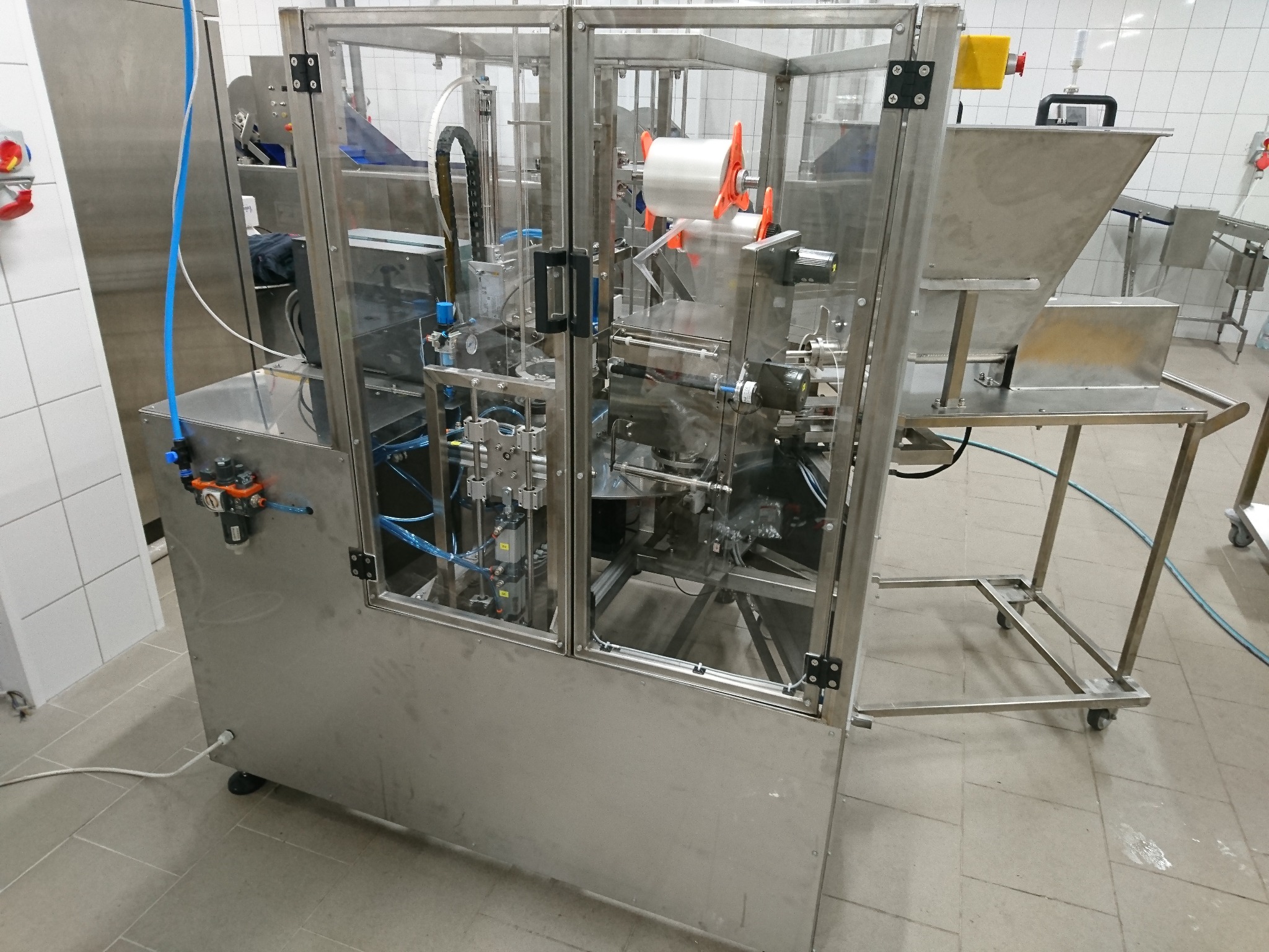 Automatyczna maszyna pakująca z przezroczystą obudową, widoczne elementy mechaniczne i rolka folii, w tle fragment linii produkcyjnej i białe kafelki.