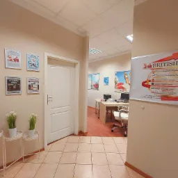 Wnętrze biura szkoły językowej z widocznymi certyfikatami i dekoracjami roślinnymi, w głębi biurka z komputerami i krzesłami.