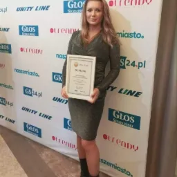 Kobieta w błyszczącej sukience i botkach pozuje z dyplomem na tle ścianki reklamowej z logo Głos Koszaliński i Trendy.