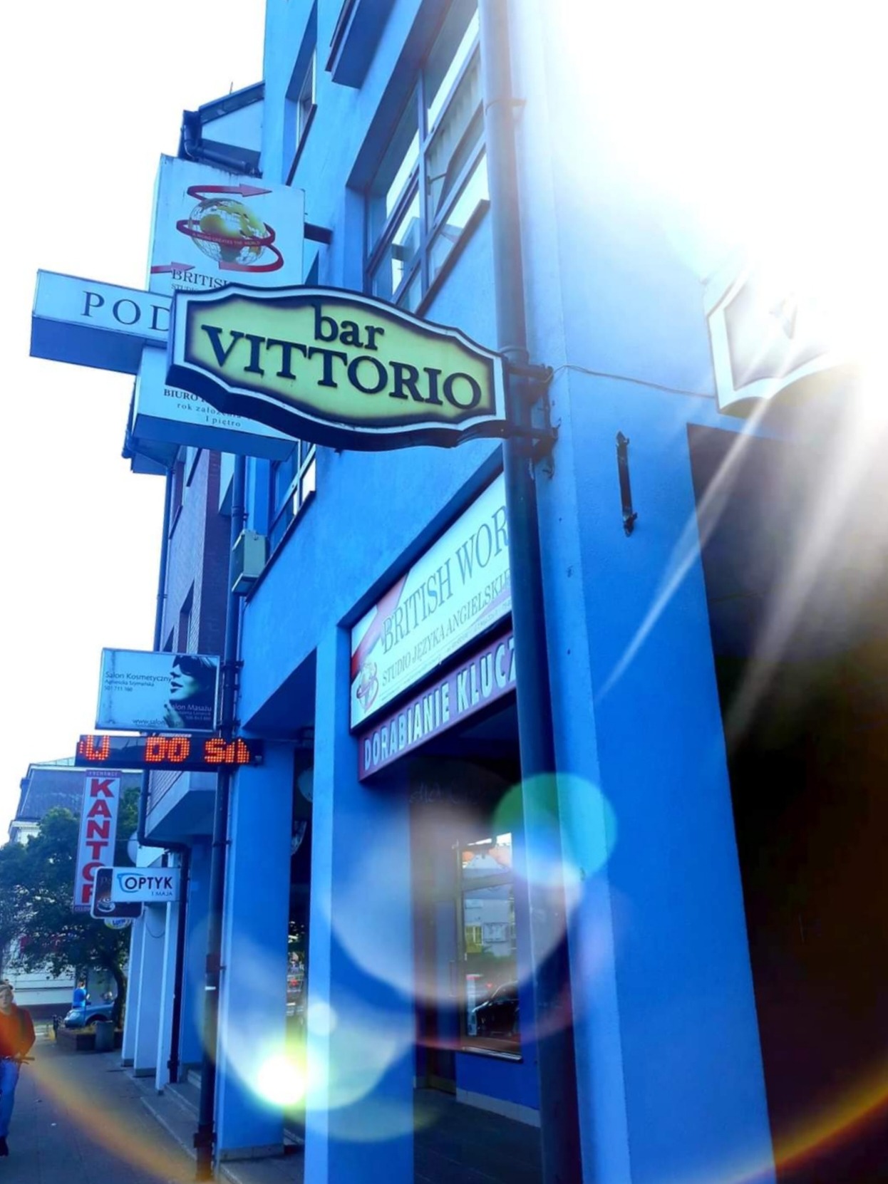 Fasada budynku w niebieskim kolorze z kilkoma szyldami, w tym 'British World Studio Języka Angielskiego' oraz 'bar Vittorio', z silnym blaskiem słońca w prawym górnym rogu.