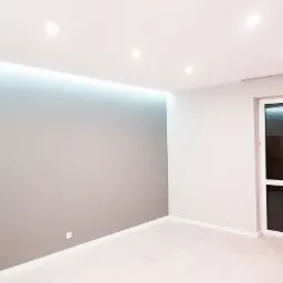 Wnętrze pokoju po remoncie, ściana pomalowana na dwa odcienie szarości, biała podłoga, sufit z oświetleniem LED i punktowym, widoczne białe drzwi balkonowe.