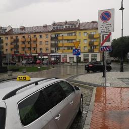 Taxi Nysa Grzegorz - Szary samochód typu kombi z żółtym oznaczeniem Taxi na dachu, zaparkowany na mokrym, brukowanym placu w Nysie, w tle budynki mieszkalne i znaki drogowe.