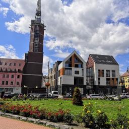 Taxi Nysa Grzegorz - Rynek w Nysie z zabytkową wieżą ciśnień, nowoczesnymi budynkami i kwitnącymi rabatami kwiatowymi w słoneczny dzień.