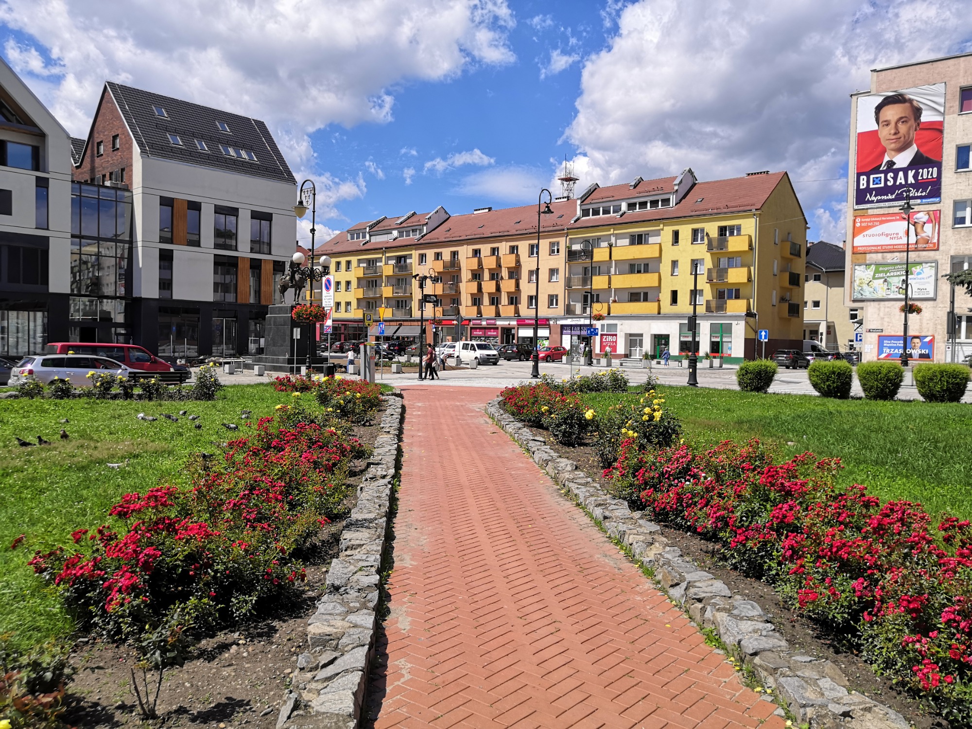 Rynek w Nysie z widokiem na budynki, chodnik z czerwonej kostki i klomby z czerwonymi różami w słoneczny dzień.