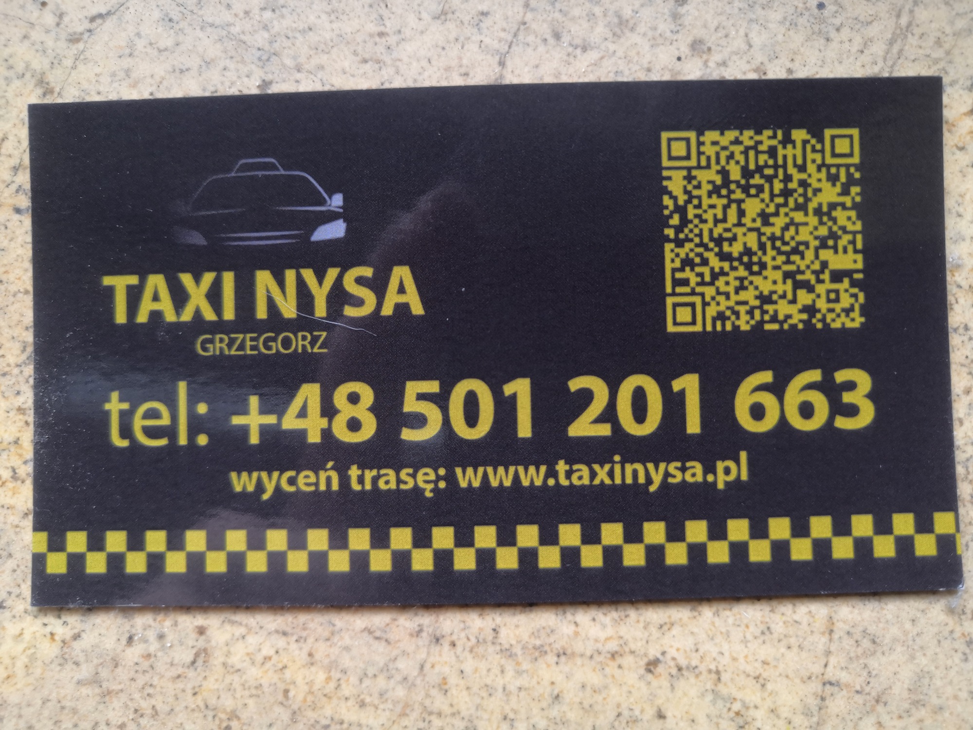 Wizytówka Taxi Nysa Grzegorz z numerem telefonu, adresem strony internetowej i kodem QR na czarnym tle z żółtymi elementami graficznymi, leżąca na beżowym podłożu.