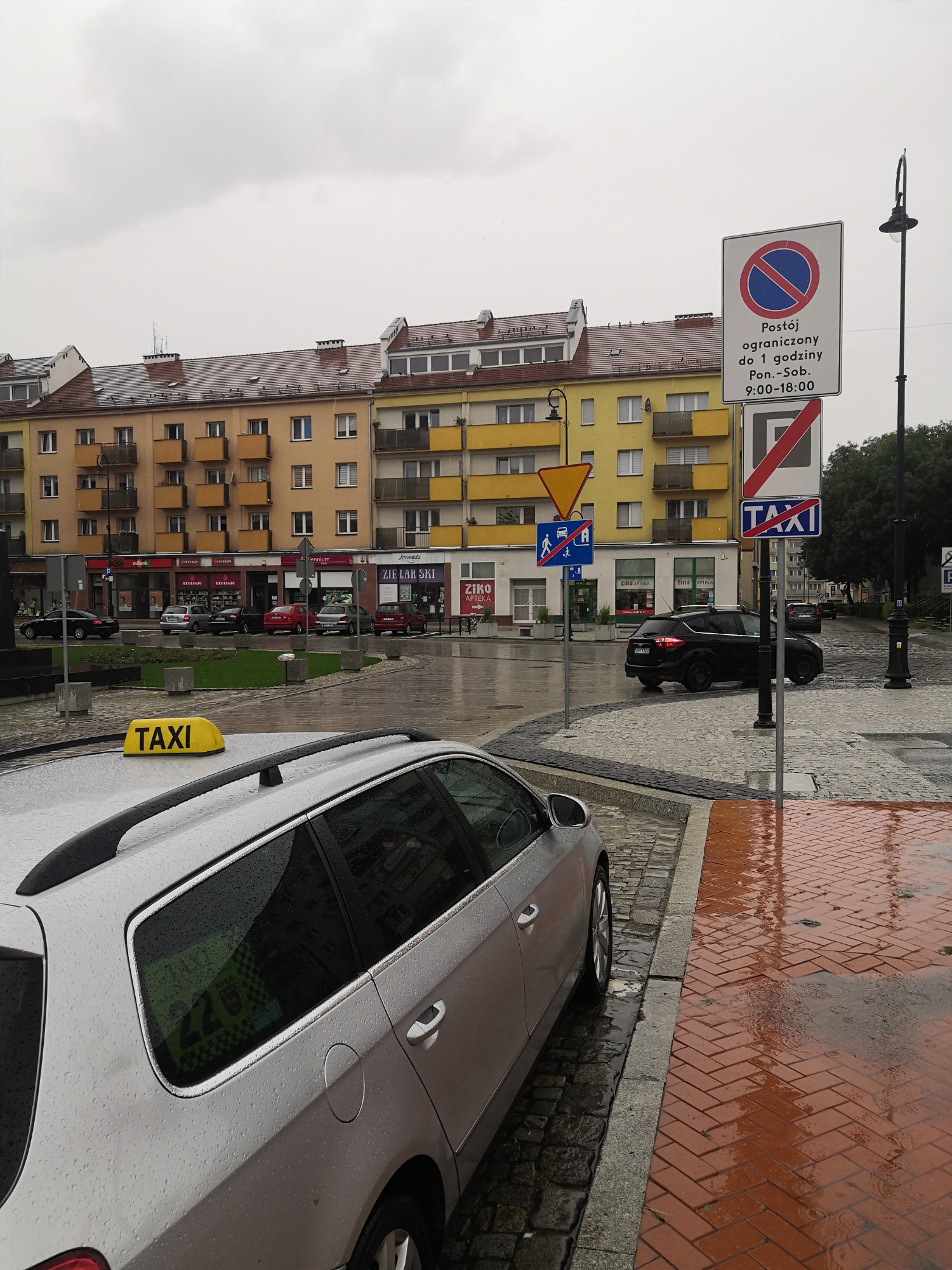 Szary samochód typu kombi z żółtym oznaczeniem Taxi na dachu, zaparkowany na mokrym, brukowanym placu w Nysie, w tle budynki mieszkalne i znaki drogowe.