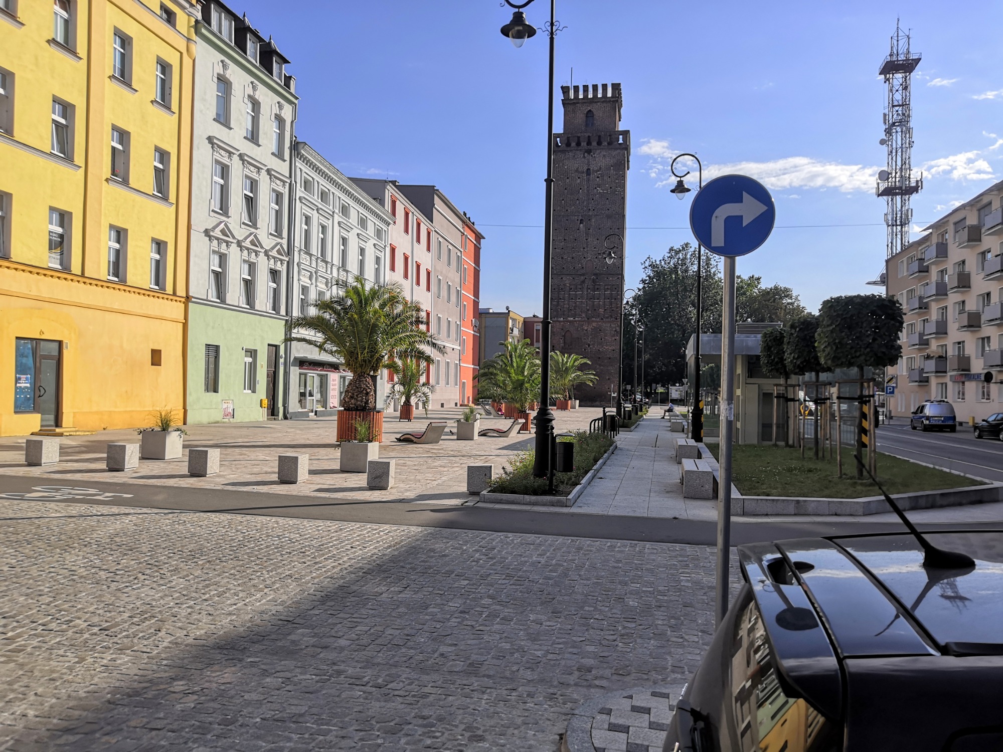 Miejski plac z kostki brukowej z palmami w donicach, ławkami i wieżą w tle, częściowo zacieniony przez zaparkowany samochód.