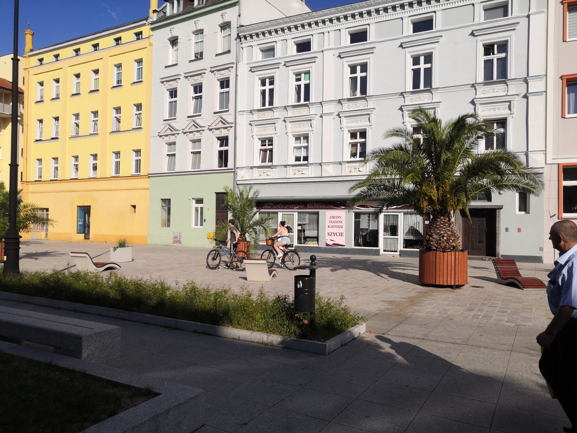 Rynek w Nysie z rowerzystami przejeżdżającymi obok kolorowych kamienic i palm w donicach, widoczny fragment mężczyzny w niebieskiej koszuli po prawej stronie.