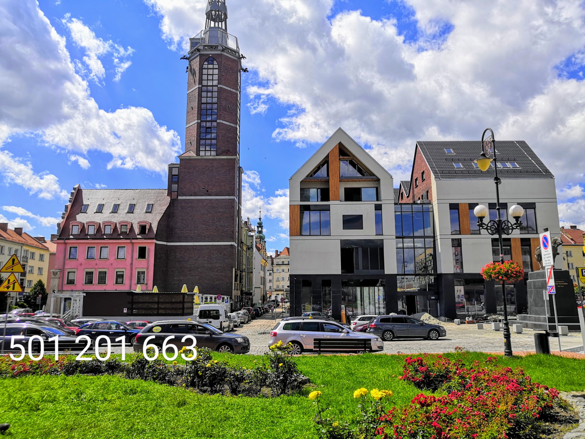 Panorama miasta Nysa z widocznymi samochodami taxi, kwiatami i charakterystyczną wieżą ratuszową w tle.