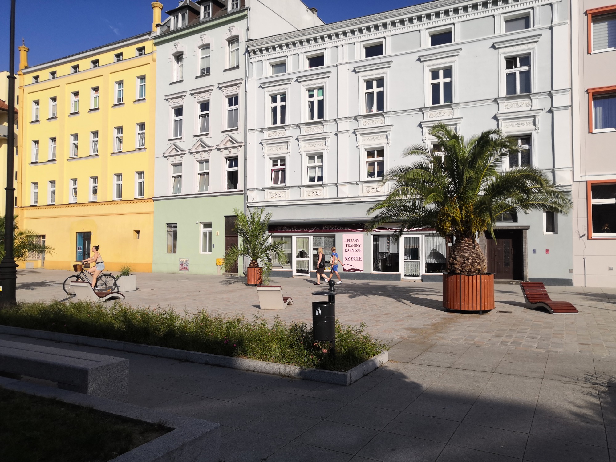 Rynek miejski w Nysie z kobietą na rowerze, palmami w donicach i dwoma osobami wchodzącymi do sklepu z szyldem 'Dekoracje Okien' oferującego firany, tkaniny, karnisze i szycie.