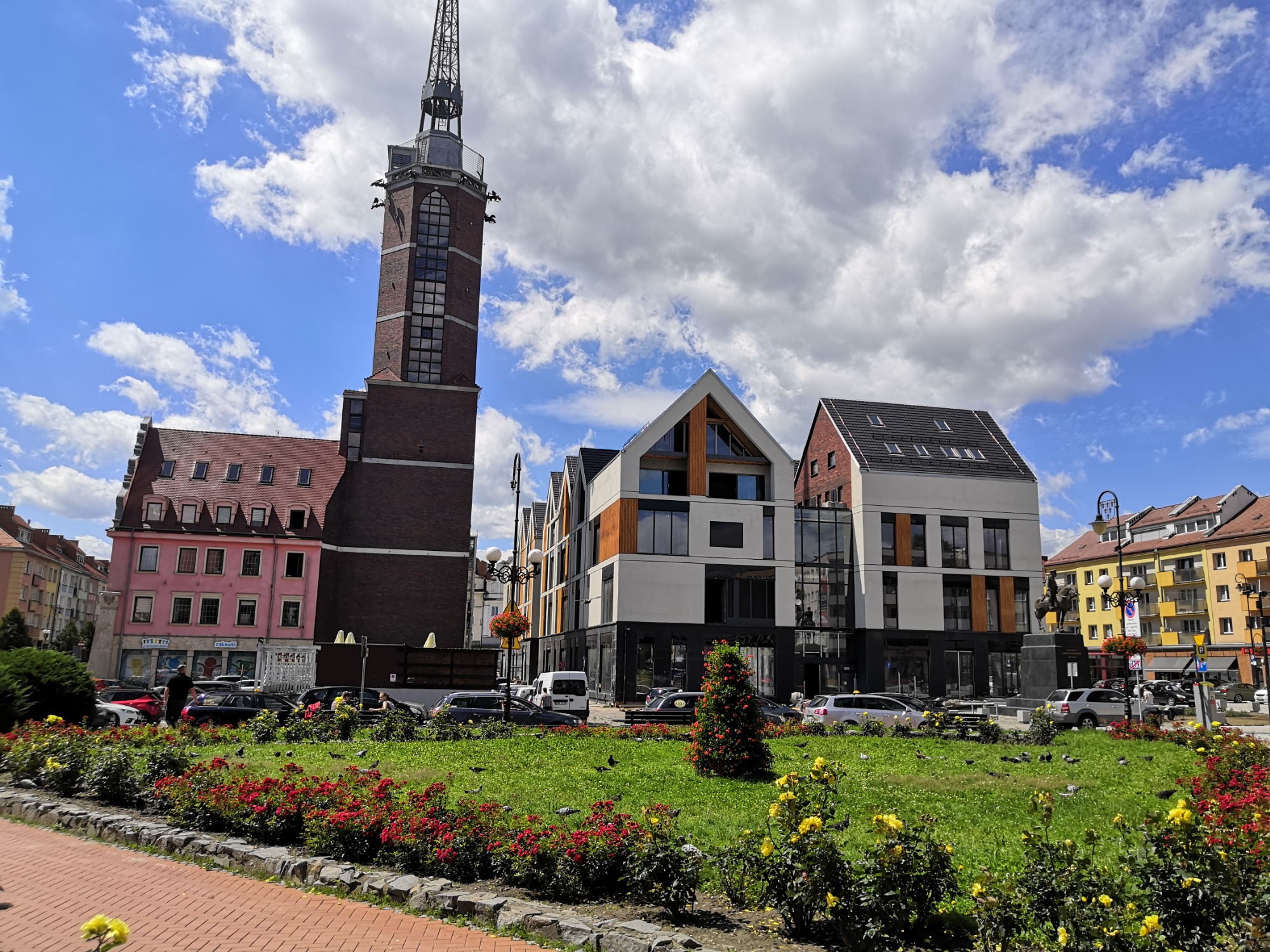 Rynek w Nysie z zabytkową wieżą ciśnień, nowoczesnymi budynkami i kwitnącymi rabatami kwiatowymi w słoneczny dzień.