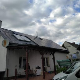 BeFe Group SC - Biały dom z ciemnym dachem i panelami słonecznymi. Widoczna weranda z drewnianymi słupami i dekoracyjnymi lampkami. Na dachu komin i antena satelitarna.