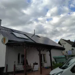 Biały dom z ciemnym dachem i panelami słonecznymi. Widoczna weranda z drewnianymi słupami i dekoracyjnymi lampkami. Na dachu komin i antena satelitarna.