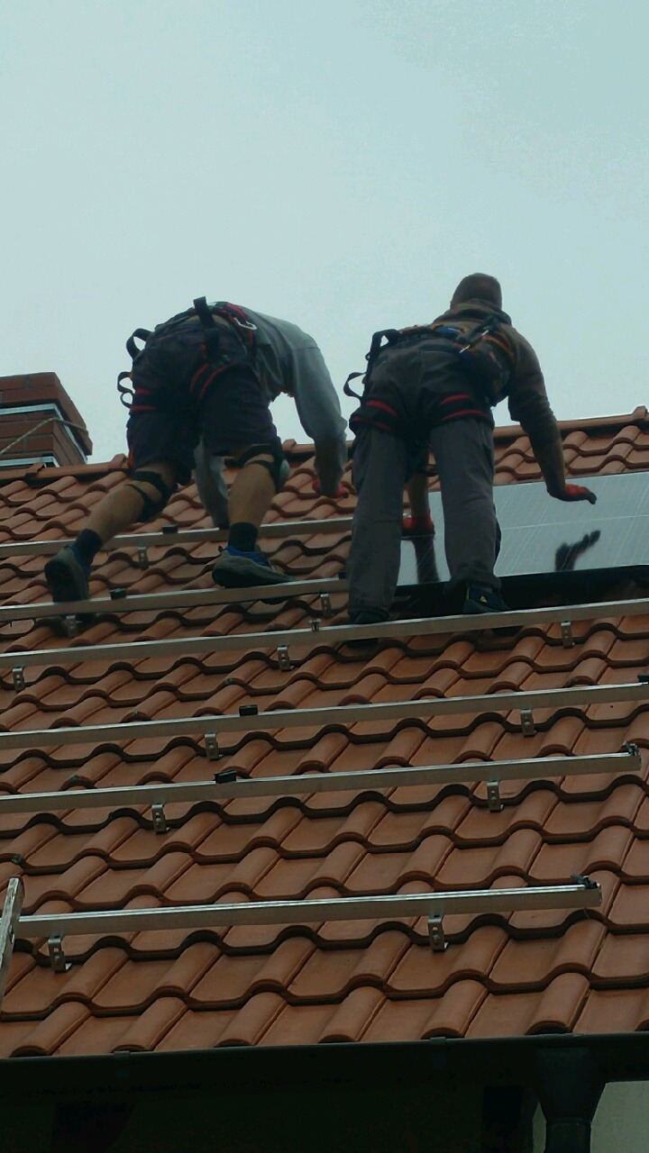 Montaż paneli fotowoltaicznych na dachu z czerwonej dachówki w Zielonej Górze. Dwóch monterów w uprzężach instaluje panele na aluminiowych szynach.