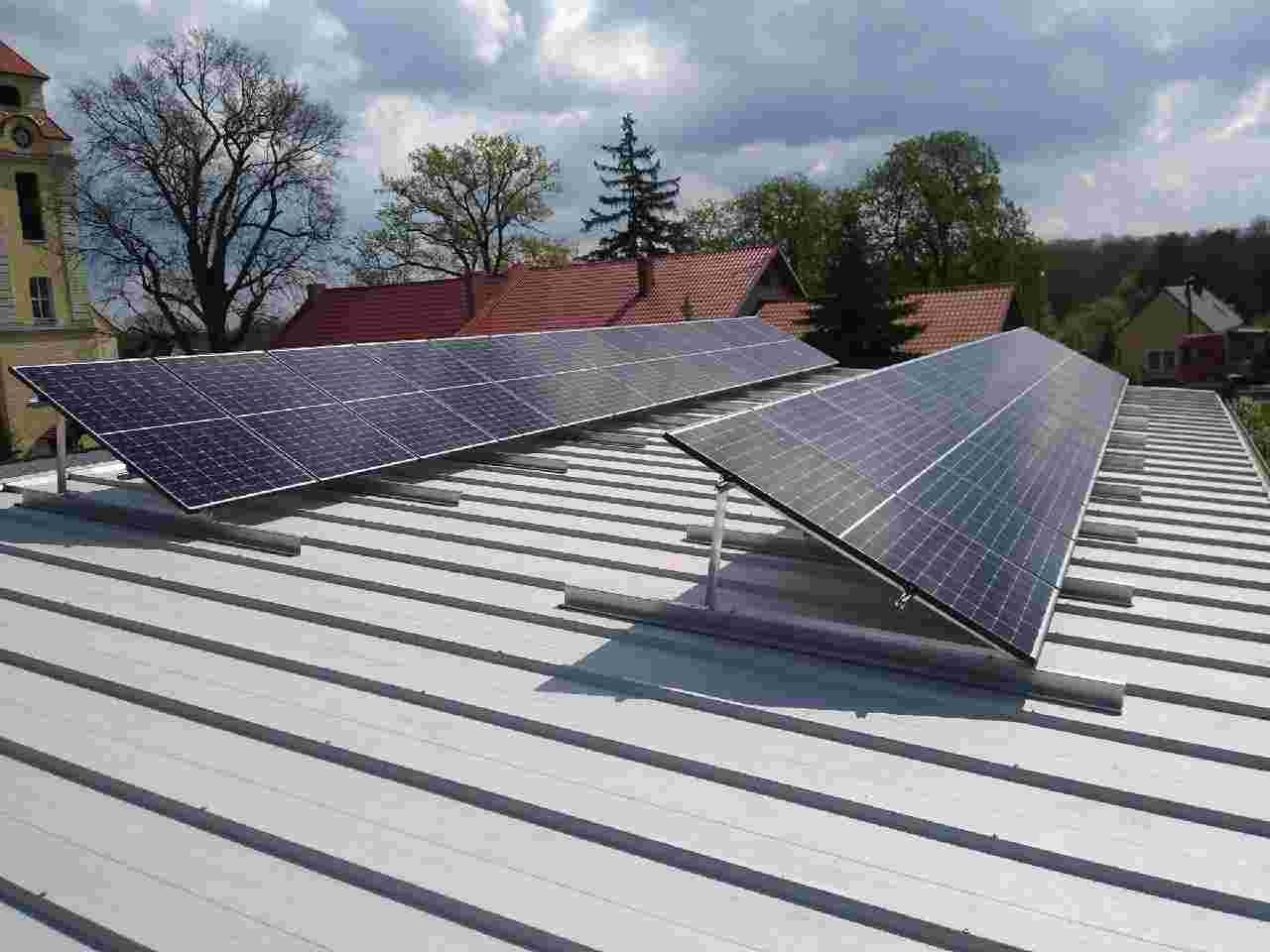 Panele fotowoltaiczne na dachu z blachy trapezowej, montowane na szarych profilach. W tle budynek z wieżyczką i drzewa pod błękitnym, pochmurnym niebem.