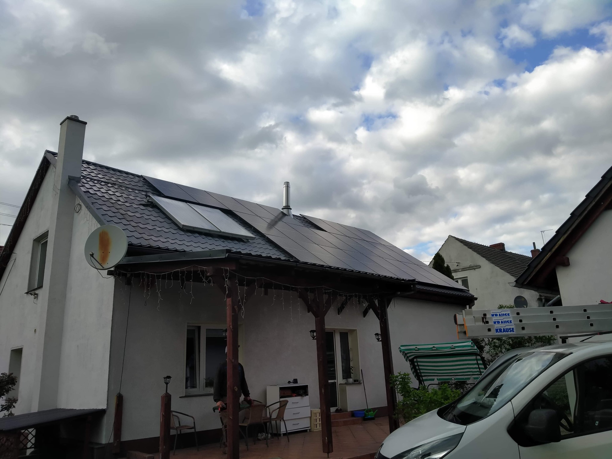 Biały dom z ciemnym dachem i panelami słonecznymi. Widoczna weranda z drewnianymi słupami i dekoracyjnymi lampkami. Na dachu komin i antena satelitarna.