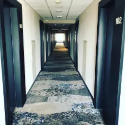 Długi korytarz hotelowy w Lublinie z wzorzystą wykładziną w odcieniach szarości i błękitu, widok w kierunku jasnego światła na końcu.