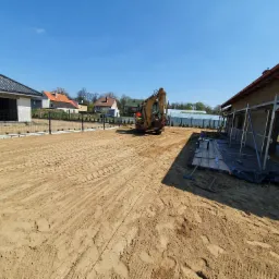 Żółta koparka na wyrównanym terenie budowy, widoczne ślady opon na piasku, w tle dom w trakcie budowy i ogrodzenie.