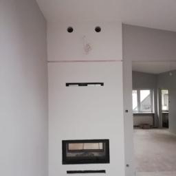 Remontypodklucz24 - Ściana z wbudowanym kominkiem w trakcie wykończenia, widoczne otwory na oświetlenie i wystające przewody, surowy stan podłogi i widok na sąsiednie pomieszczenie.