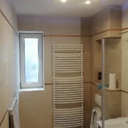 Wykończona łazienka z beżowymi płytkami, podwieszanym sufitem z oświetleniem LED, grzejnikiem drabinkowym i częściowo widoczną toaletą oraz umywalką z szafką, na podłodze tuba silikonu.