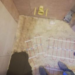 Remontypodklucz24 - Montaż ogrzewania podłogowego elektrycznego w łazience, widoczne kable grzewcze DEVI na podłożu z naniesioną zaprawą klejową, w tle rury kanalizacyjne wystające ze ściany.