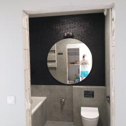 Remontypodklucz24 - Wnętrze łazienki w trakcie prac wykończeniowych, widoczne szare płytki, czarna mozaika, biała armatura i okrągłe lustro z odbiciem osoby robiącej zdjęcie, surowy ościeżnica drzwi wskazuje...