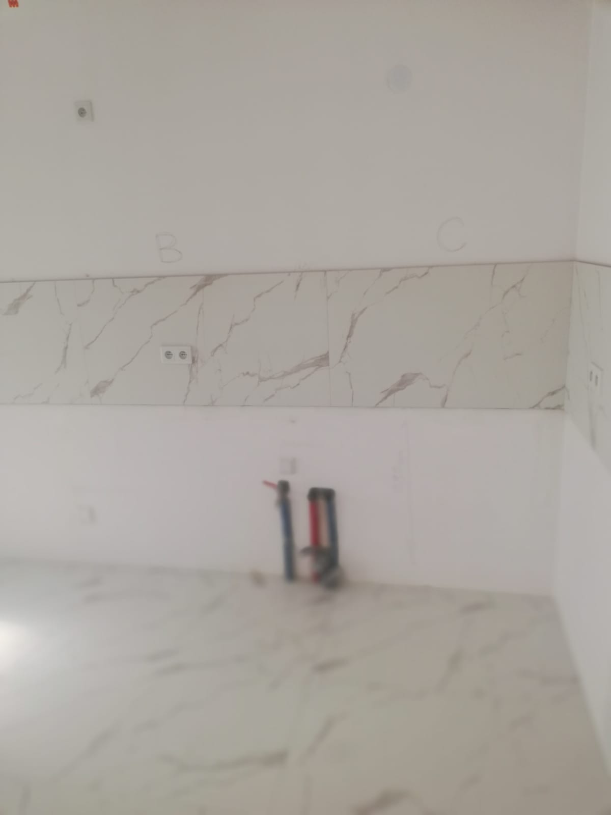 Niedokończona łazienka z ułożonymi płytkami imitującymi marmur na ścianie, widoczne instalacje wodne, częściowo zamazane tło i napisy 'B' oraz 'C' na ścianie.