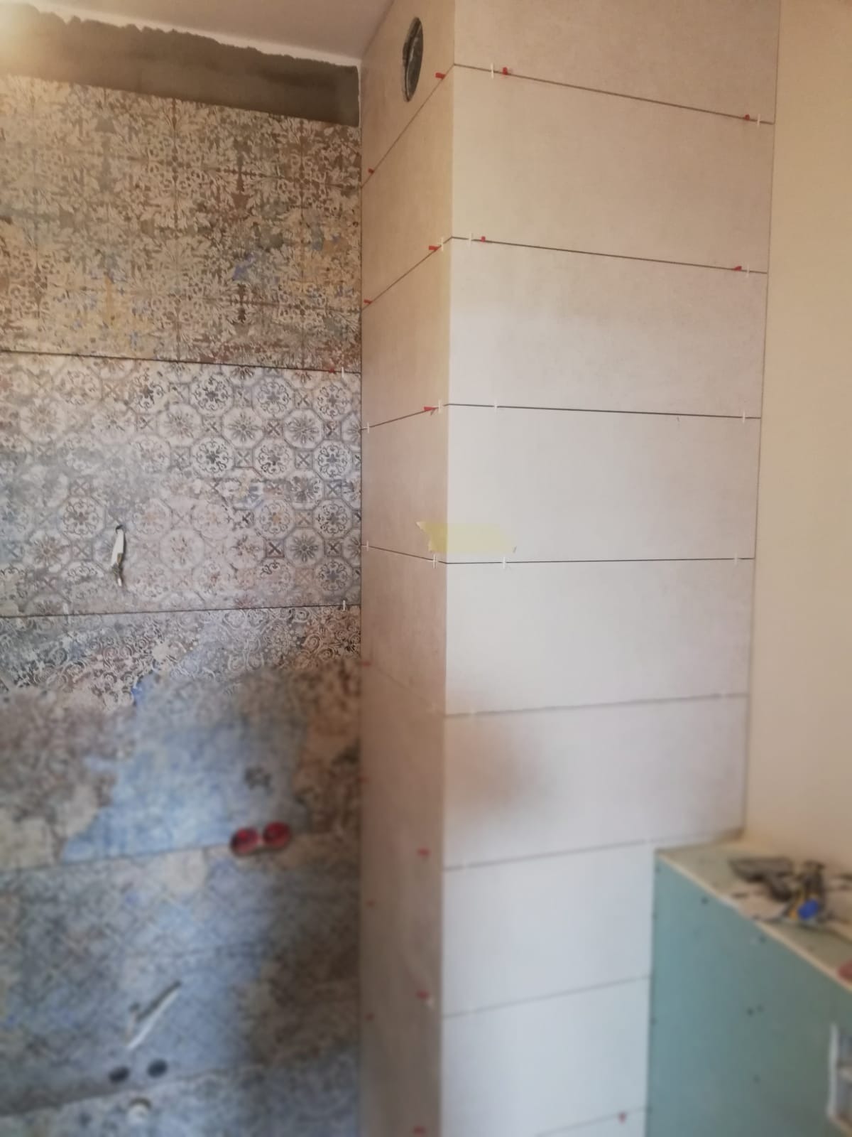 Ściana łazienki w trakcie układania płytek: część ściany wykończona płytkami wzorzystymi, obok pionowy element z jasnych, prostokątnych płytek, z widocznymi krzyżykami dystansowymi i taśmą maskującą.