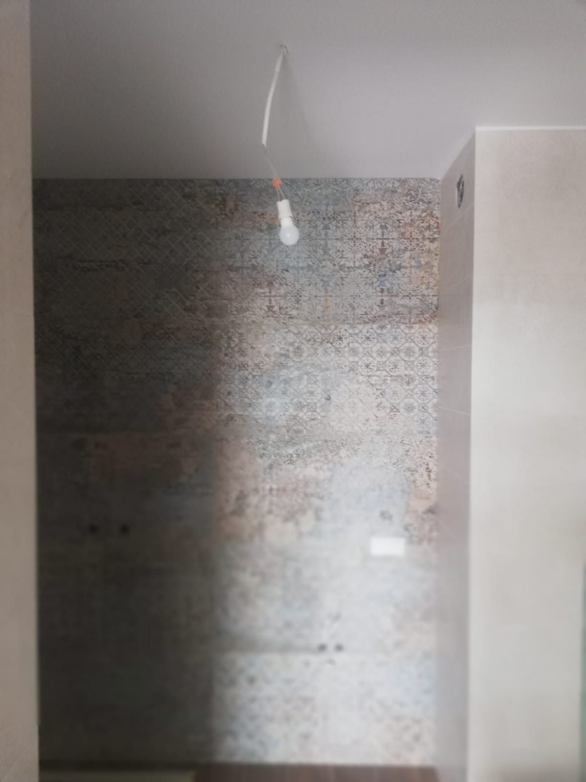 Ściana łazienki w trakcie wykańczania, z widocznymi płytkami o wzorzystym, postarzanym wyglądzie i wiszącą żarówką na kablu.