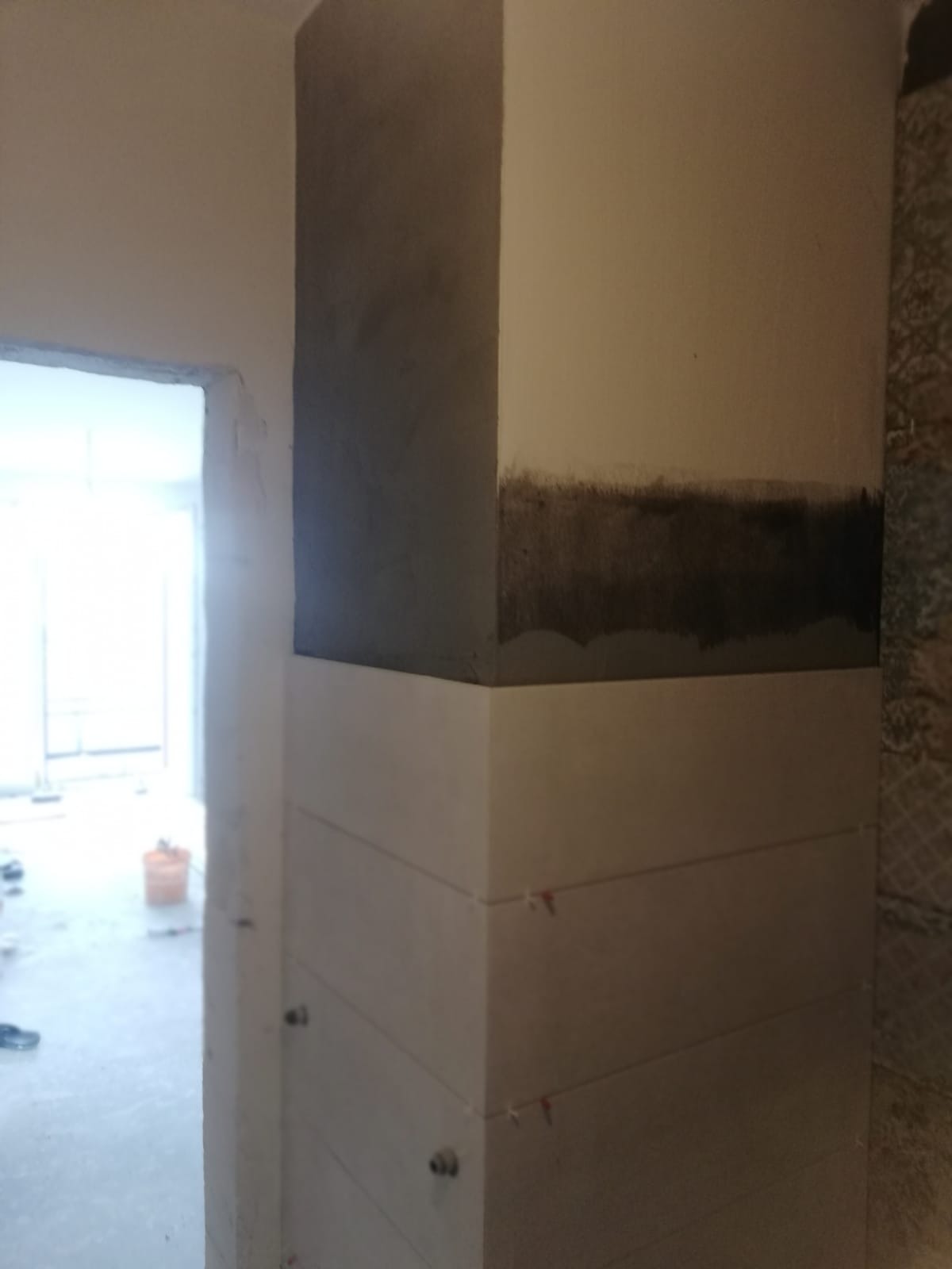 Wewnętrzny narożnik ściany z częściowo położonymi płytkami, widoczne krzyżyki dystansowe i ślady po gruntowaniu, w tle widoczne pomieszczenie w trakcie prac.