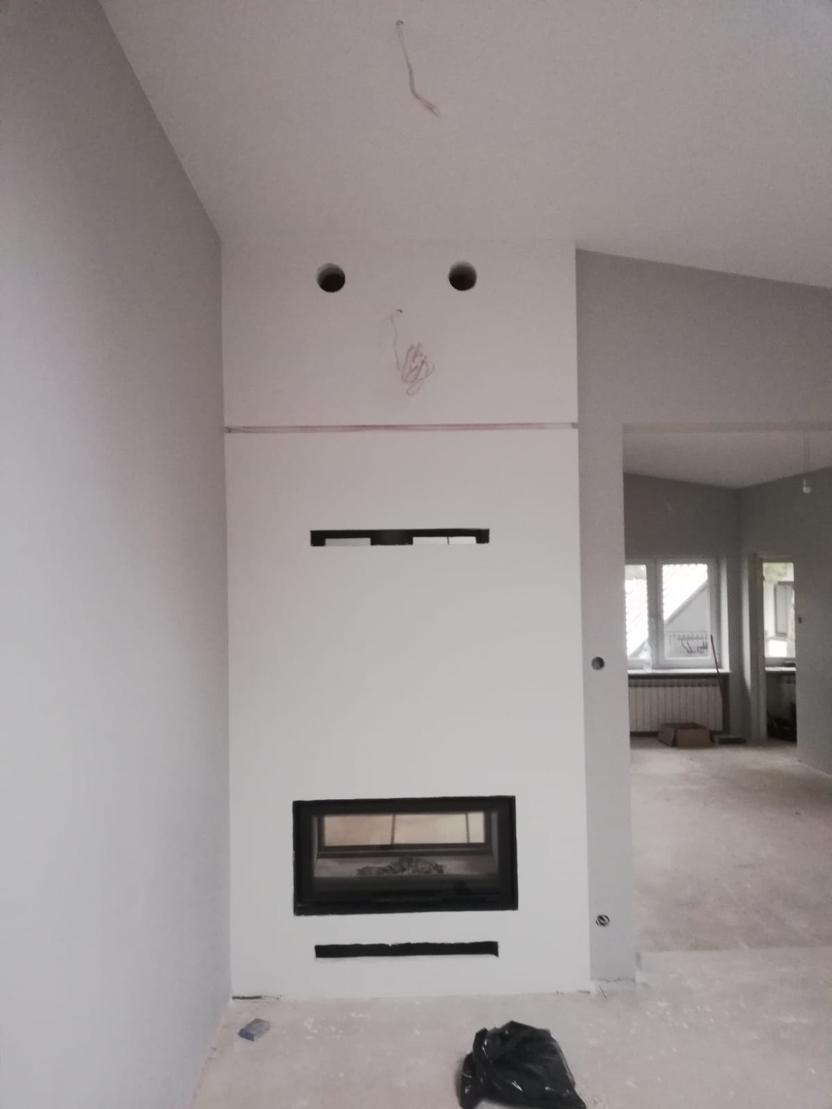 Ściana z wbudowanym kominkiem w trakcie wykończenia, widoczne otwory na oświetlenie i wystające przewody, surowy stan podłogi i widok na sąsiednie pomieszczenie.