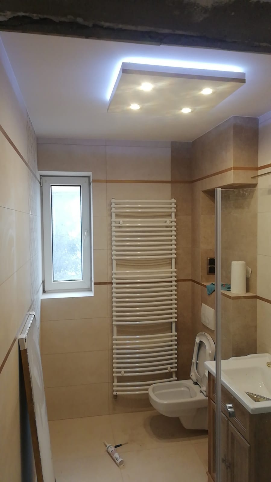 Wykończona łazienka z beżowymi płytkami, podwieszanym sufitem z oświetleniem LED, grzejnikiem drabinkowym i częściowo widoczną toaletą oraz umywalką z szafką, na podłodze tuba silikonu.