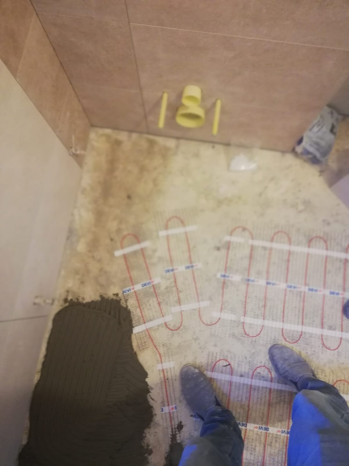 Montaż ogrzewania podłogowego elektrycznego w łazience, widoczne kable grzewcze DEVI na podłożu z naniesioną zaprawą klejową, w tle rury kanalizacyjne wystające ze ściany.