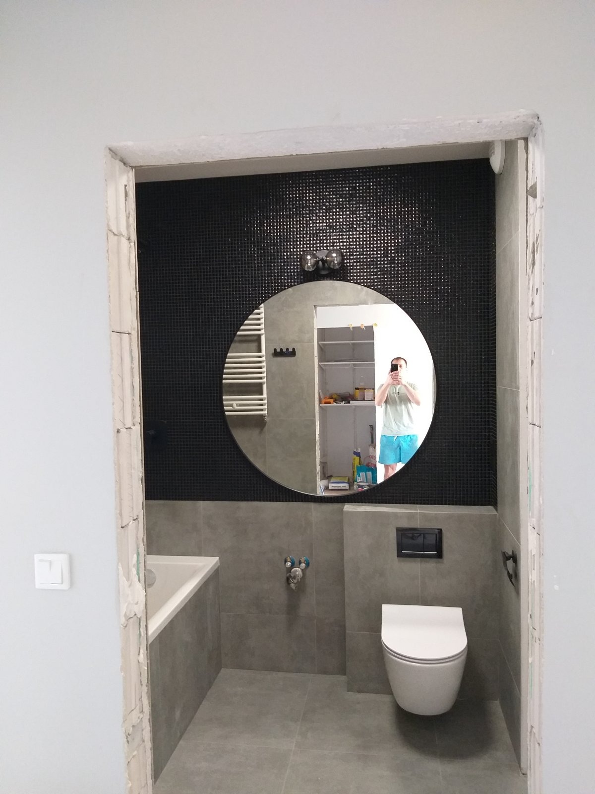 Wnętrze łazienki w trakcie prac wykończeniowych, widoczne szare płytki, czarna mozaika, biała armatura i okrągłe lustro z odbiciem osoby robiącej zdjęcie, surowy ościeżnica drzwi wskazuje...