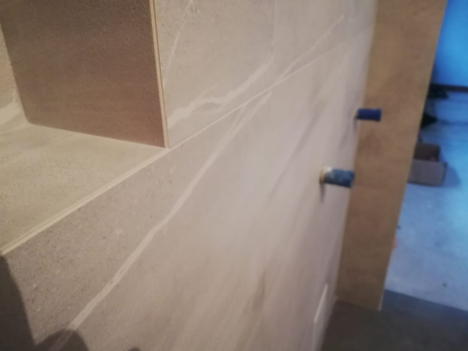 Ściana wyłożona jasnymi płytkami imitującymi marmur z wnęką i wystającymi niebieskimi końcówkami rur instalacyjnych.