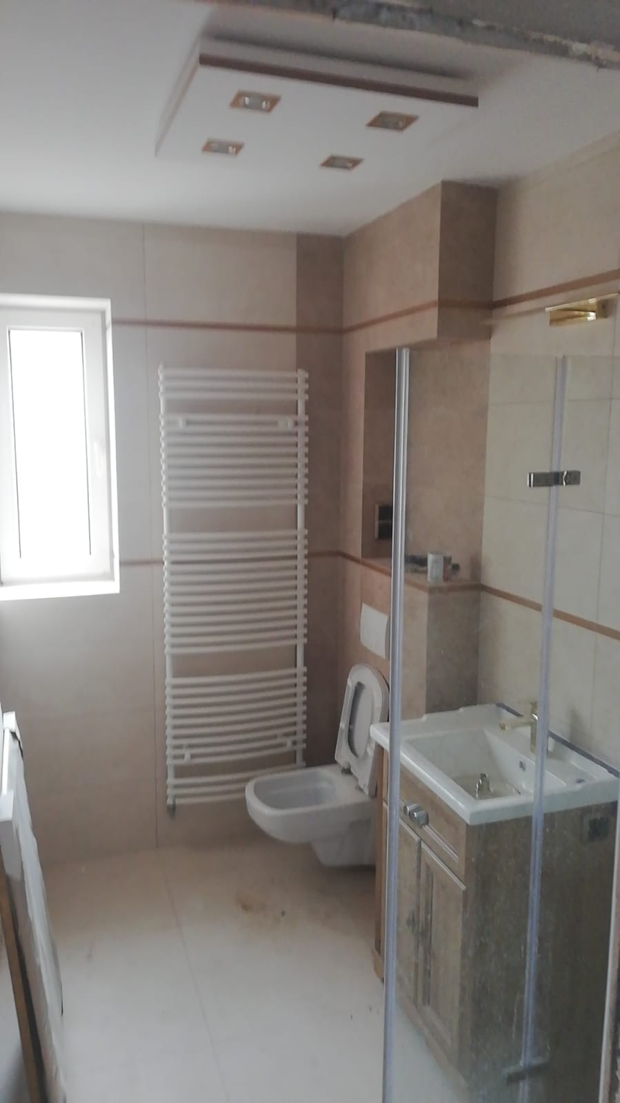 Wnętrze łazienki w trakcie prac wykończeniowych, widoczna biała toaleta, grzejnik drabinkowy, umywalka z szafką w stylu retro i częściowo zamontowana kabina prysznicowa, beżowe płytki na ścianach...