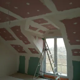 Poddasze w trakcie remontu: widoczne płyty karton gips na skośnym suficie i ścianach, częściowo zaszpachlowane łączenia, drabina aluminiowa oparta o okno, przygotowane otwory pod oświetlenie.