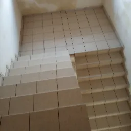 Schody wyłożone beżowymi płytkami ceramicznymi, widok z góry, ściany częściowo przygotowane pod malowanie, widoczne niedoskonałości na powierzchni.