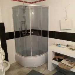 Narożna kabina prysznicowa z chromowanymi elementami w łazience z biało-czarnymi płytkami, widoczne akcesoria do montażu na blacie umywalki i szare płytki podłogowe.