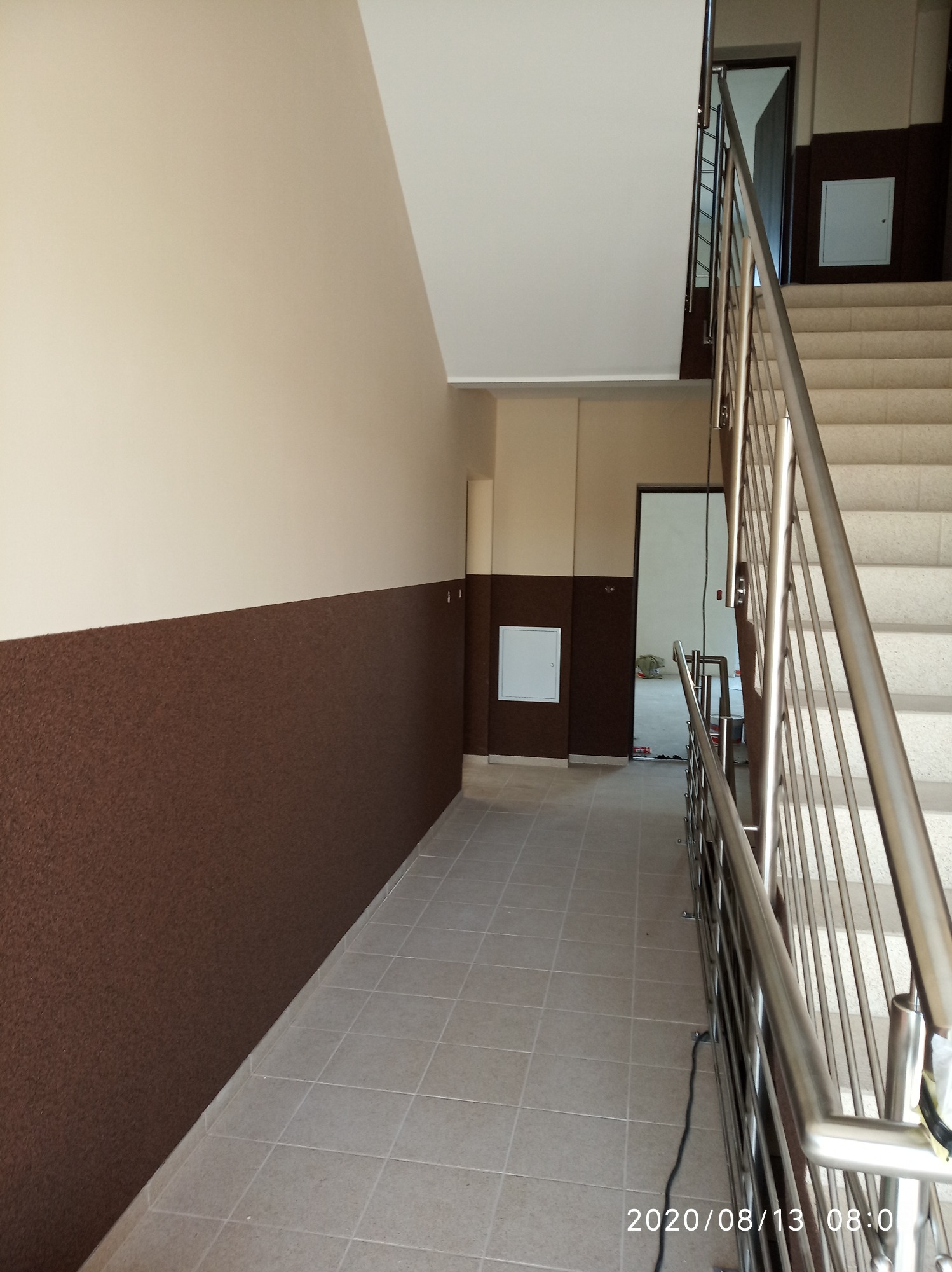 Klatka schodowa z beżowymi ścianami, brązowym pasem boazerii, szarymi płytkami na podłodze i metalową balustradą, widok z dołu schodów.