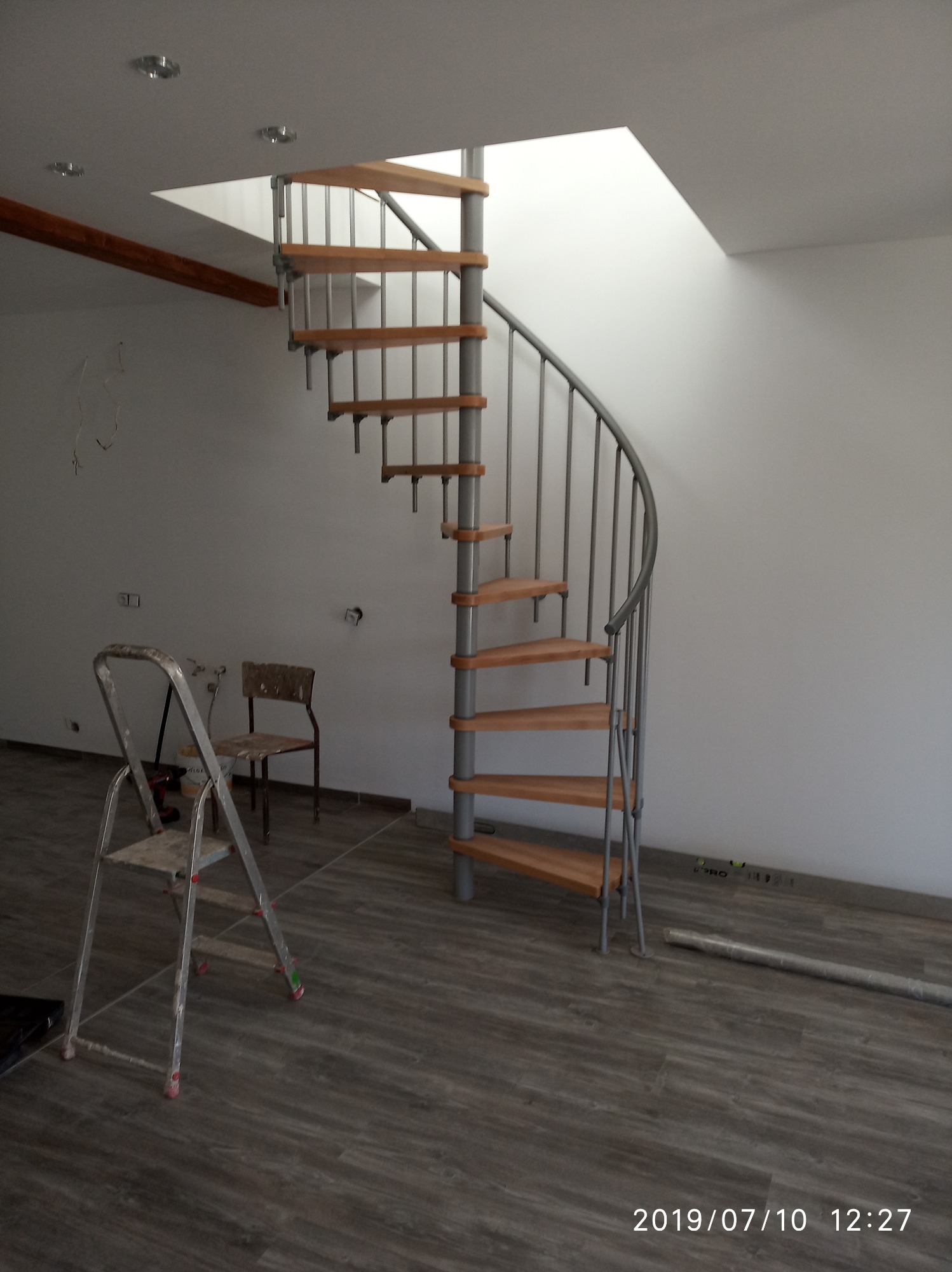 Montaż schodów spiralnych z drewnianymi stopniami i metalową konstrukcją w surowym wnętrzu, widoczna drabina malarska i poziomica przy ścianie.