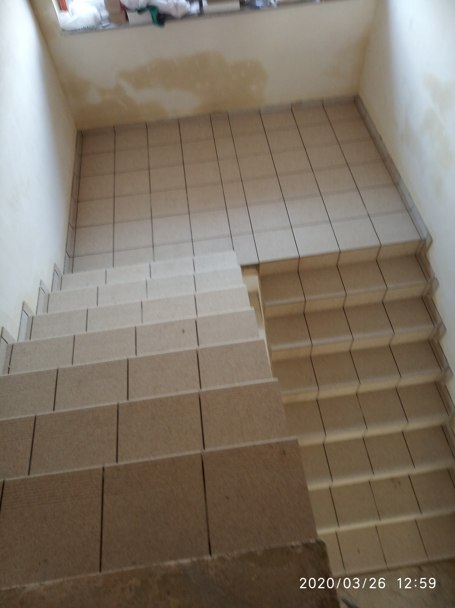 Schody wyłożone beżowymi płytkami ceramicznymi, widok z góry, ściany częściowo przygotowane pod malowanie, widoczne niedoskonałości na powierzchni.