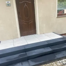 Nowe, trzypoziomowe schody wejściowe z ciemnoszarej kostki brukowej z jasnymi płytkami na górnym poziomie, prowadzące do brązowych drzwi wejściowych domu z numerem 91, obok okna i małego drzewka.