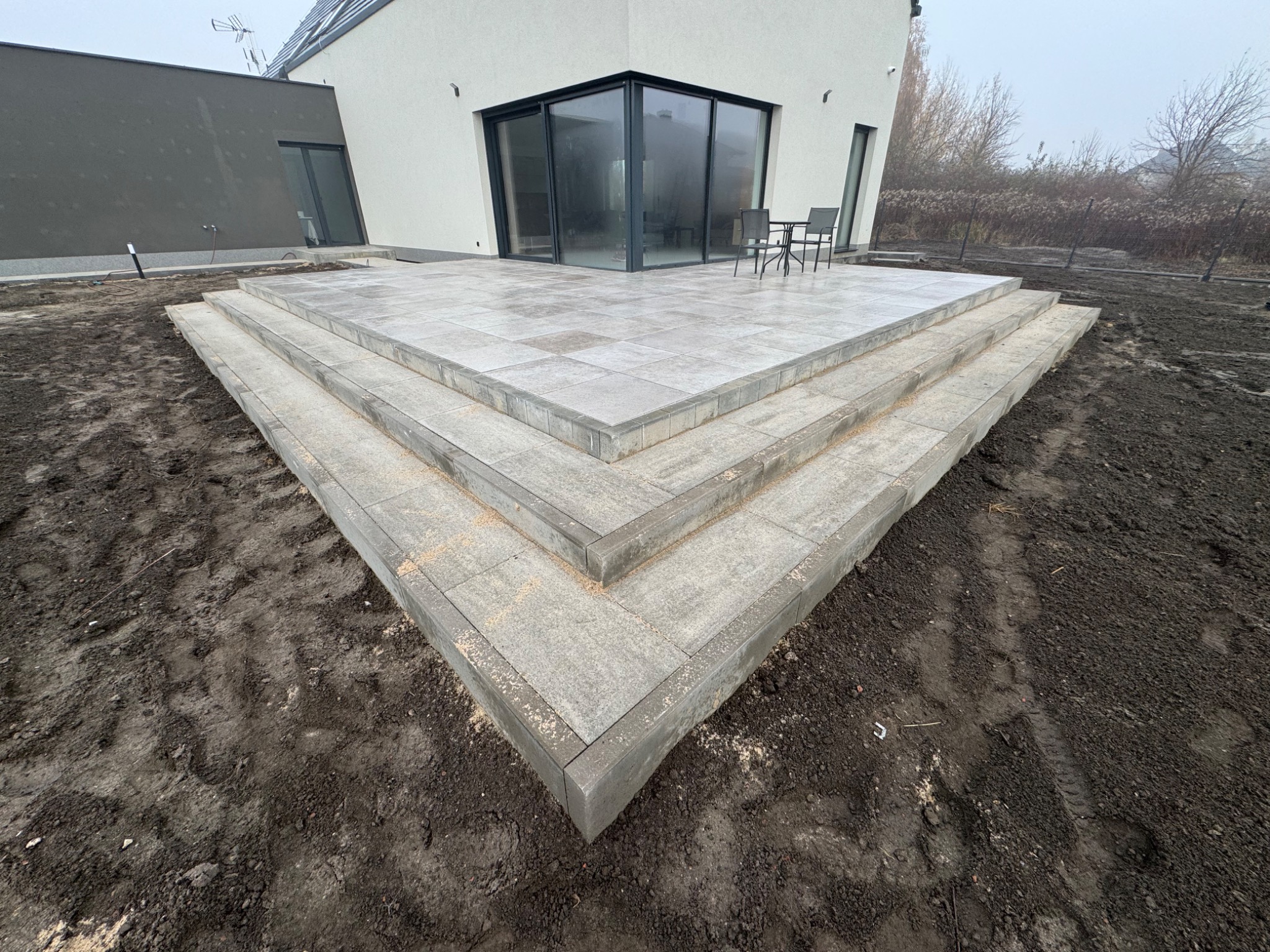 Nowoczesny taras z szarej kostki brukowej, ułożony schodkowo, prowadzący do przeszklonego salonu domu. Widoczne krzesła i stół na tarasie, ziemia wokół.