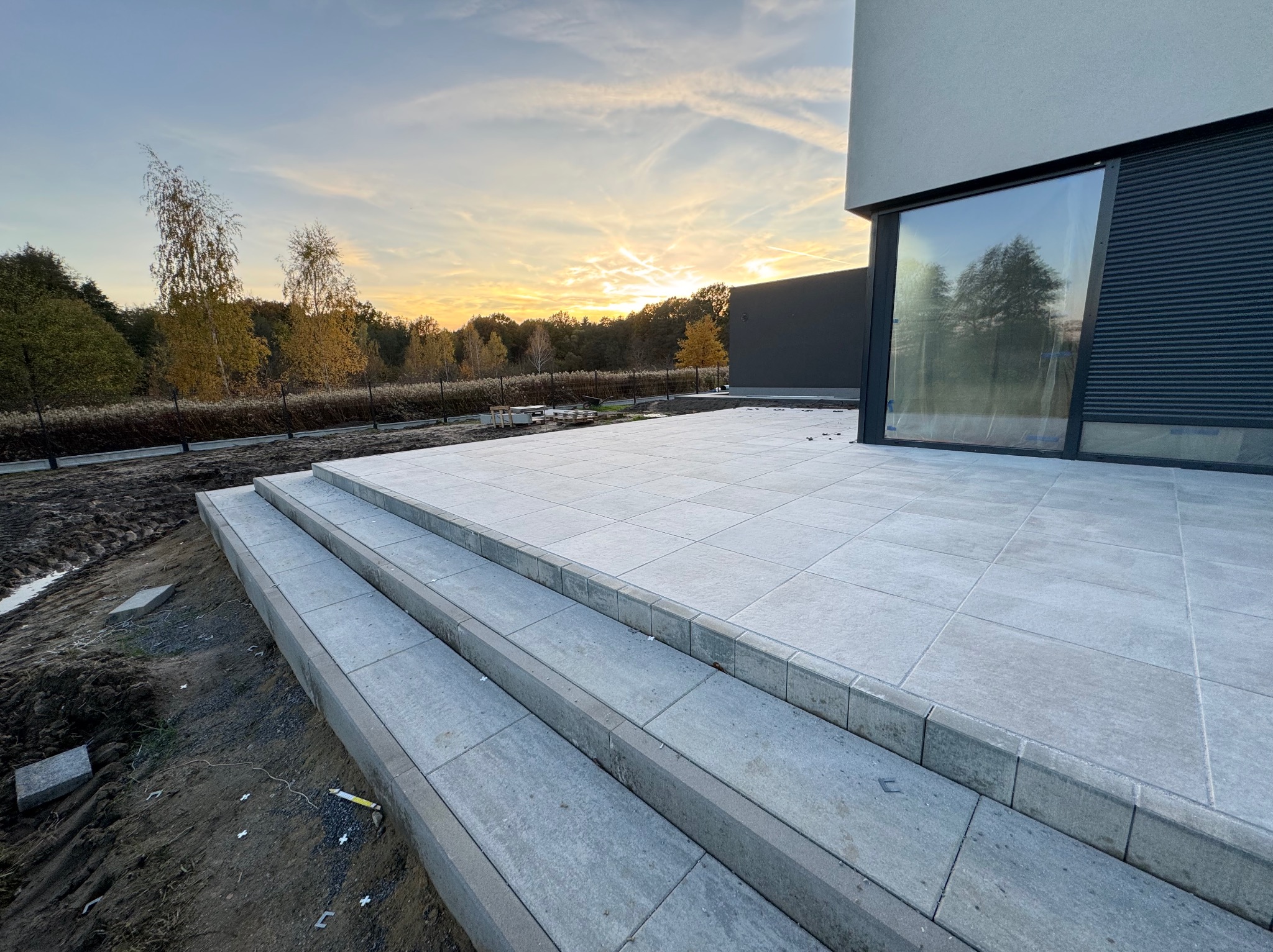 Nowoczesny taras z szarej kostki brukowej, schody prowadzące do budynku. Widok na zachodzące słońce i jesienne drzewa w tle. Minimalistyczny design.