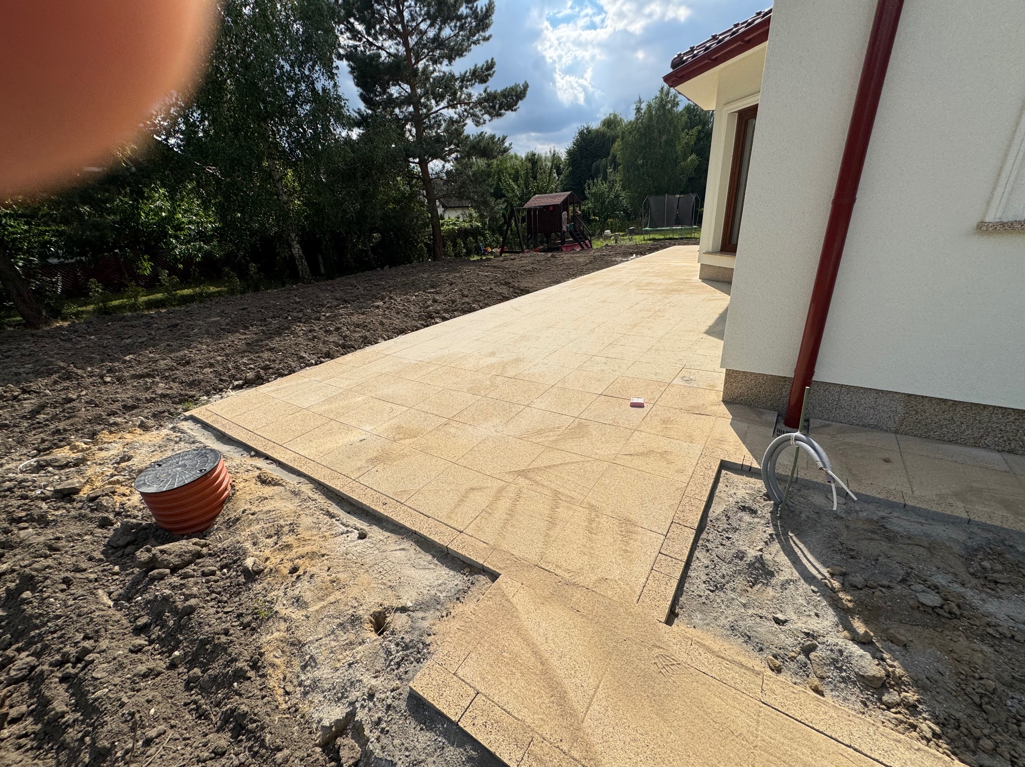 Układanie jasnej kostki brukowej na posesji domu jednorodzinnego. Widoczny fragment ułożonej nawierzchni, przygotowane podłoże i studzienka kanalizacyjna.