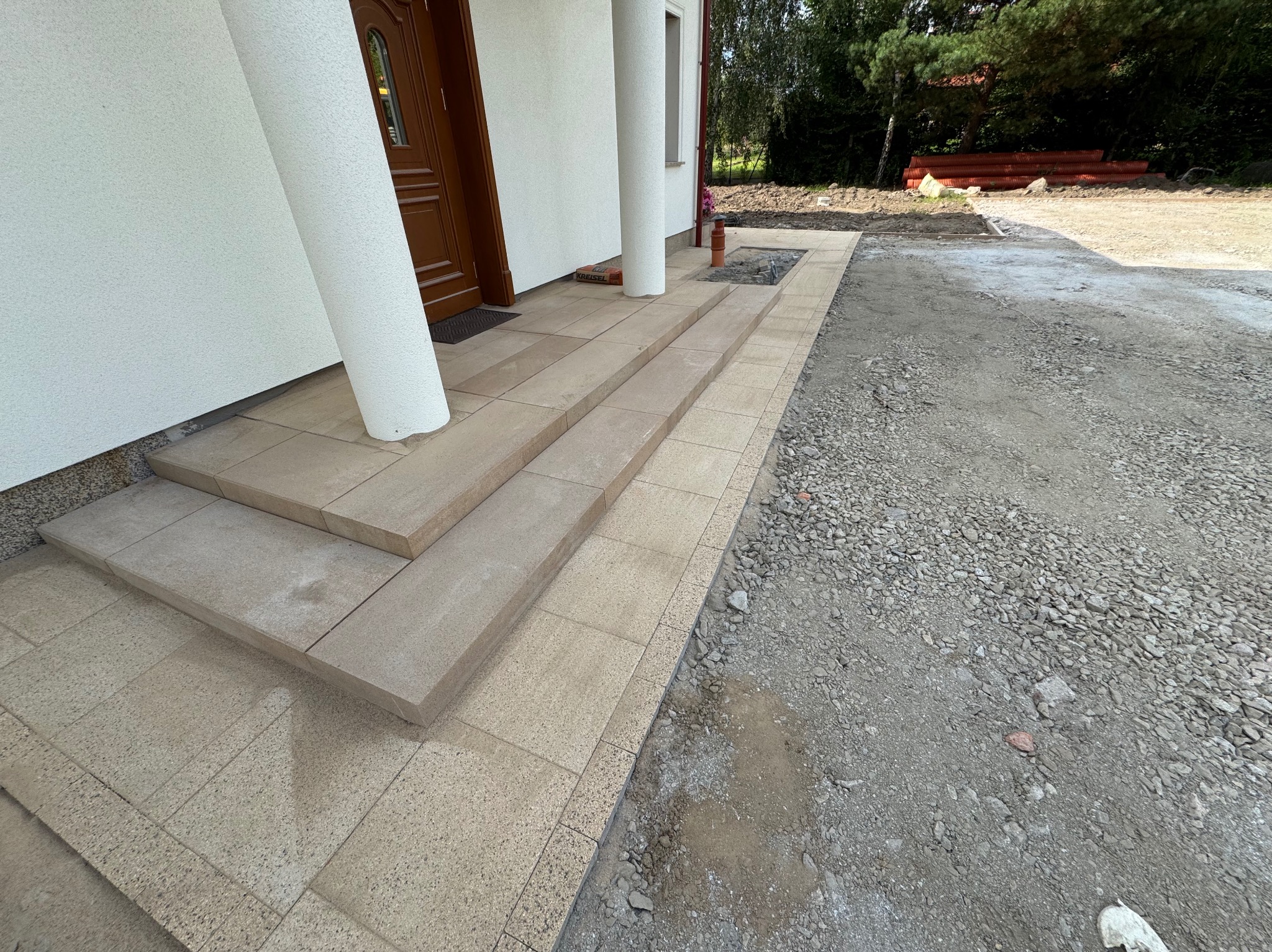 Ułożone schody i chodnik z jasnej kostki brukowej prowadzące do wejścia domu z białymi kolumnami. W tle widoczne rury i teren przygotowany pod dalsze prace.