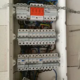 Rozdzielnica elektryczna w trakcie montażu, widoczne zabezpieczenia przepięciowe Simtec, bezpieczniki Hager, okablowanie w kolorach niebieskim, brązowym i żółto-zielonym, zamocowana do ściany...