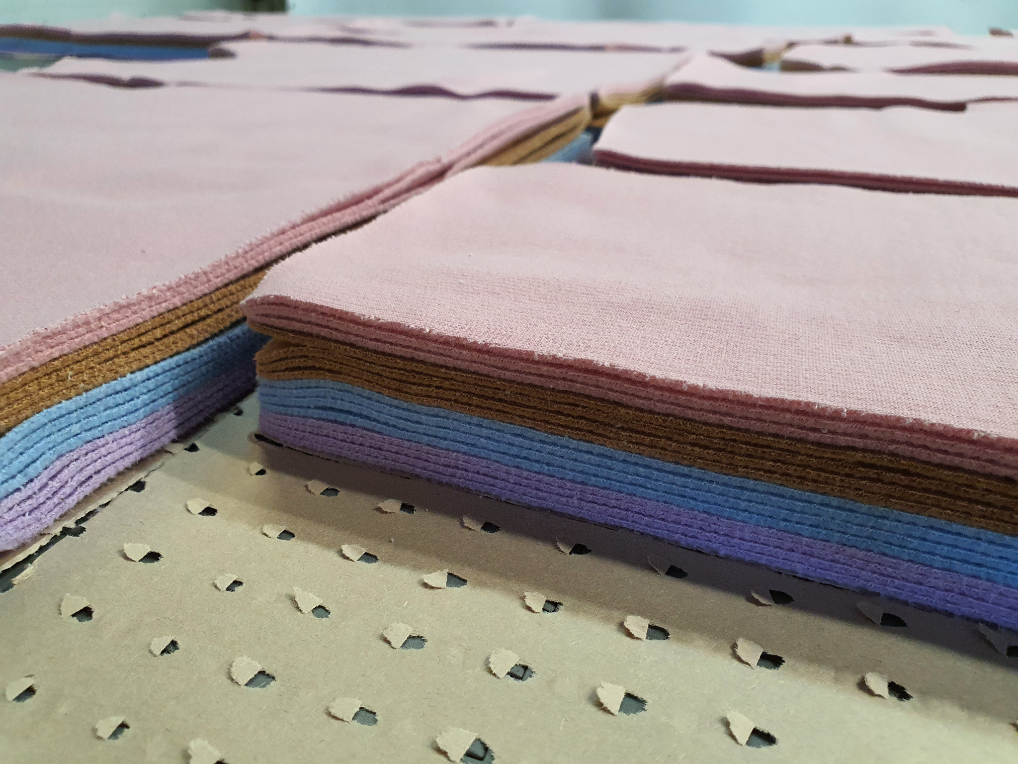 Stosy pociętej, kolorowej dresówki na perforowanym podłożu, widoczne warstwy fioletowe, niebieskie, brązowe i różowe.